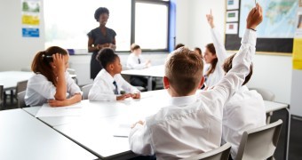 Primary School Students Wearing Uniform Raising Hands To Answer Question Set By Teacher In Classroom