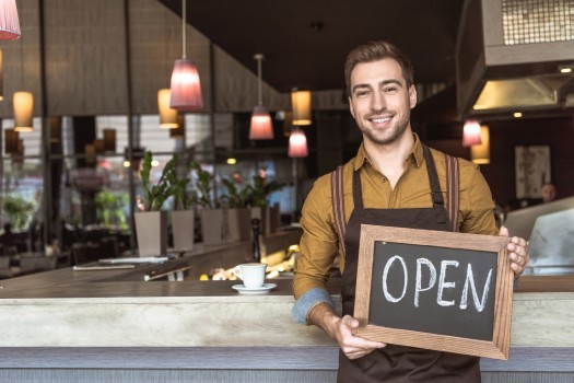 happy young waiter holding chalkboard with open inscription in cafe